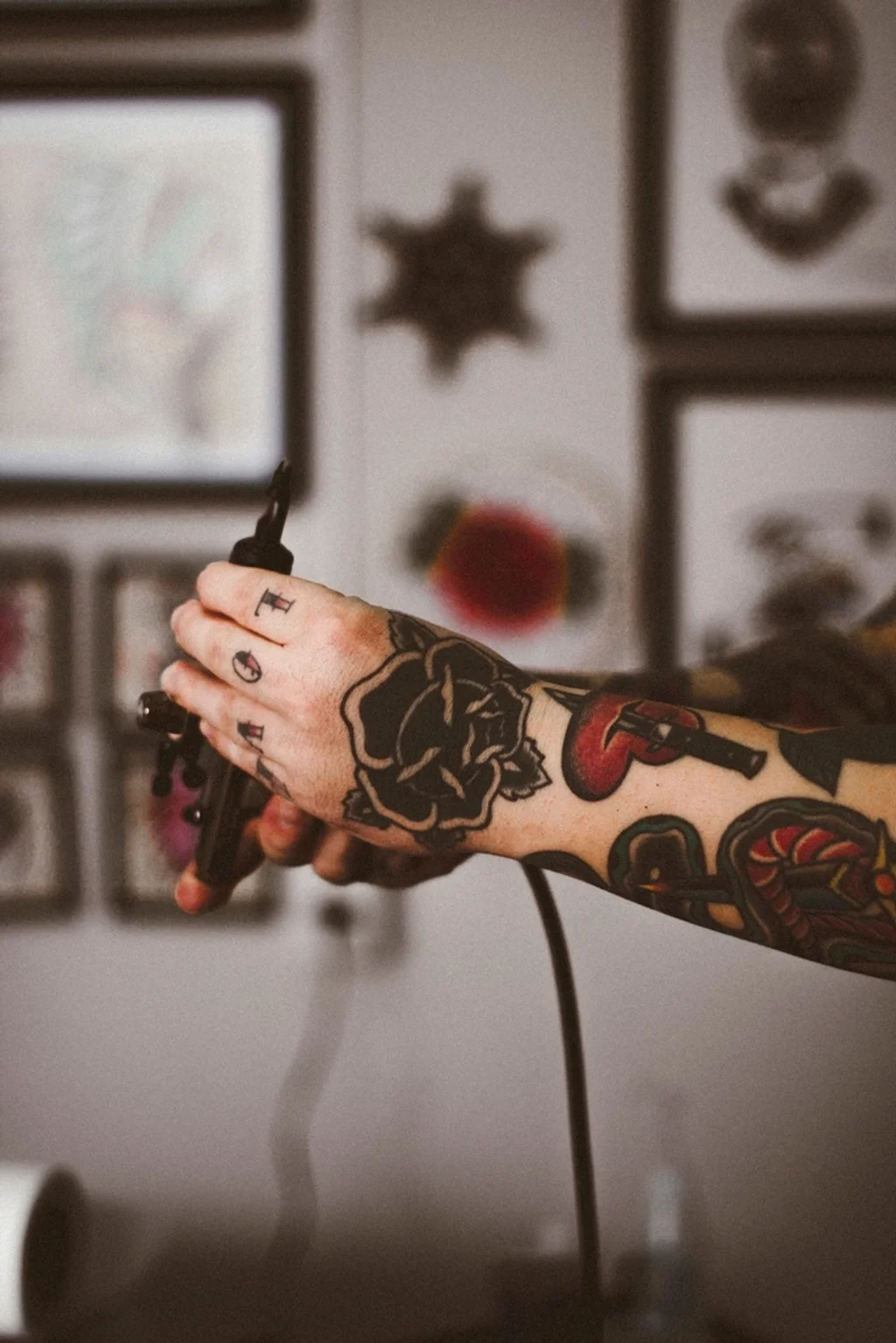 A tattooed hand holding a coil machine in front of traditional flash